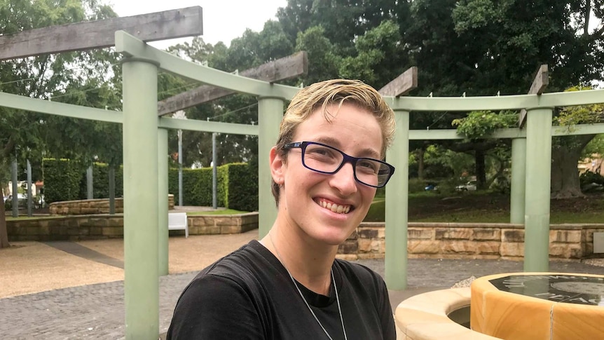 Sarah Lucock, with glasses and a black t-shirt, smiles while standing in a park.