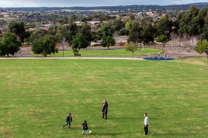 A drone shot shows a young family kicking a soccer ball on a lush green field