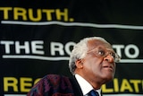 Archbishop Desmond Tutu looks thoughtful in a brown suit with the word 'truth' behind him on black sign