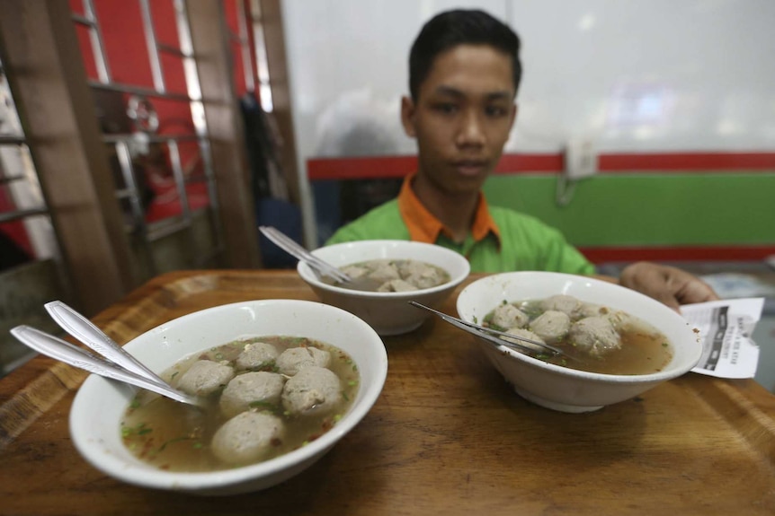 Three bowls of soup with meat dumplings on a wooden tray held by a young Indonesian man.