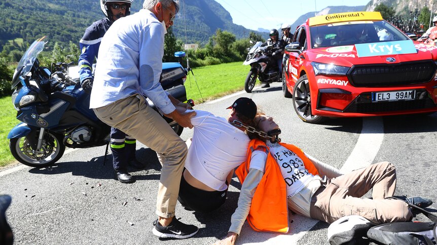 Two men sit cross-legged on a road, a thick sliver chain around their necks. A man is trying to pull one man up