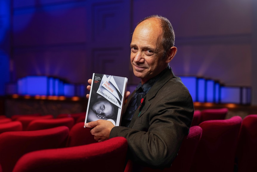 Interior photo of a 57-year-old bald man sitting in a theater with red chairs holding a copy of his book The Promise.