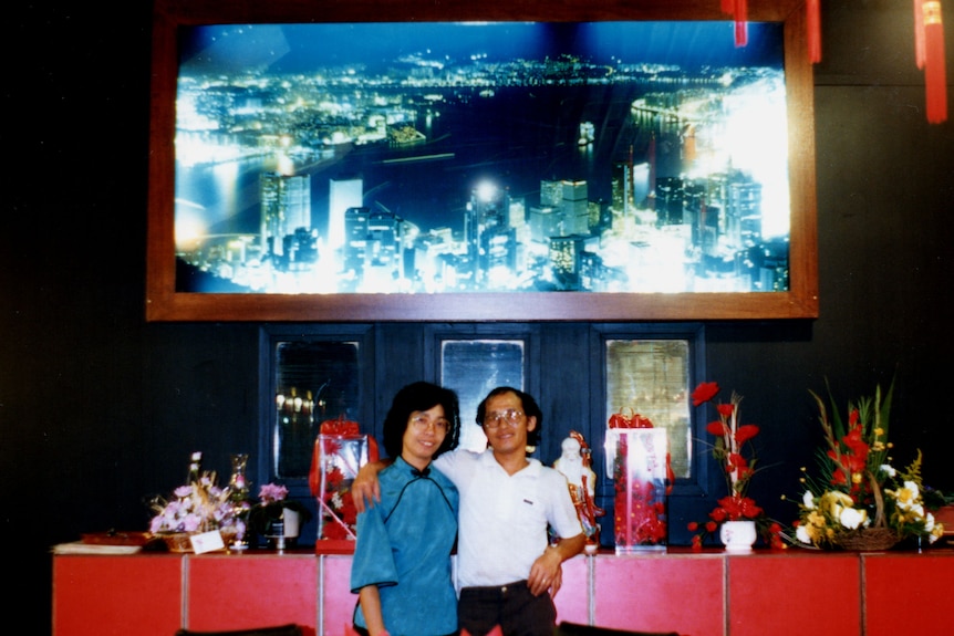 A couple standing in front of a restaurant wall with a decorative photo of Hong Kong in the background.
