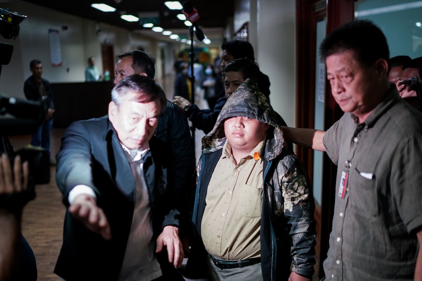 A young man with round face, wearing a beige shirt and camouflage jacket, is escorted as cameras flash nearby
