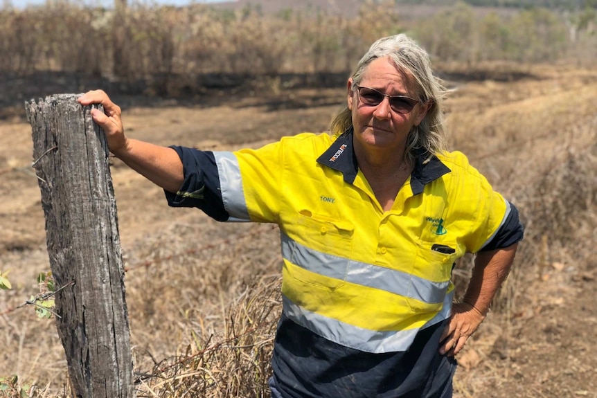 A woman in a high-vis shirt leans on the fence post of a property which, in the background appears to be burnt and fire-damaged