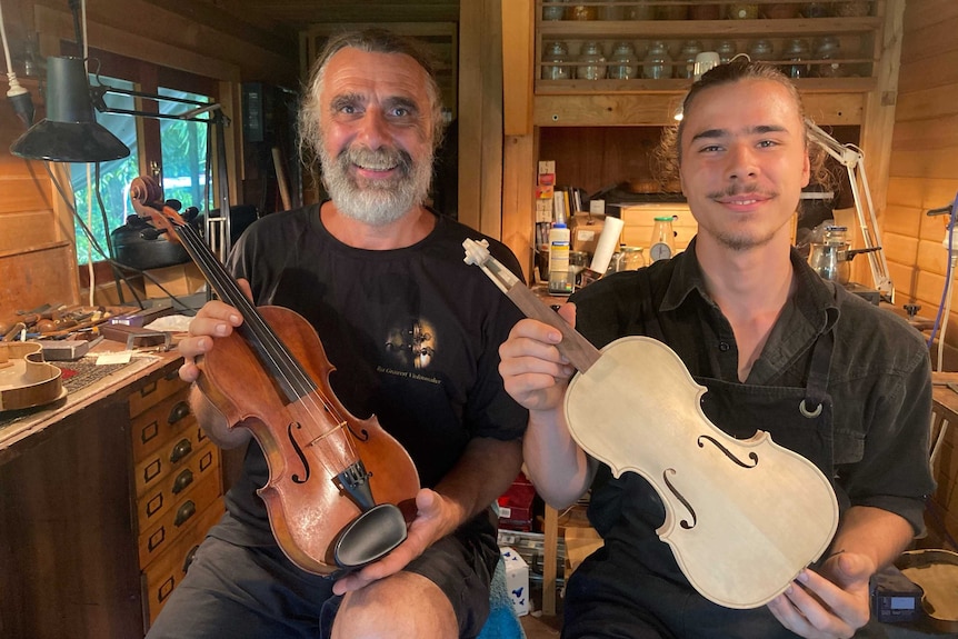 Two men, each holding a violin, sitting in their mobile workshop