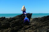 A woman on a rock wearing a mermaid tail, there is ocean in the background.
