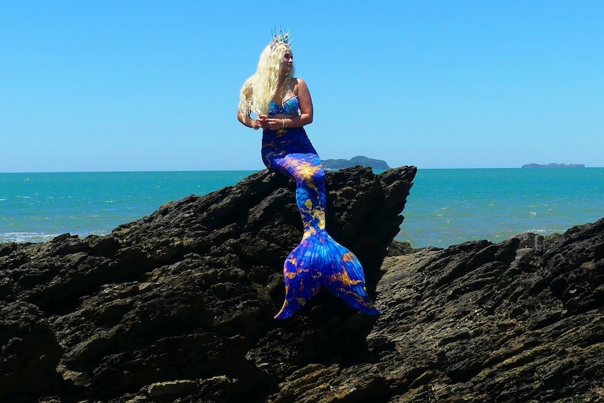 Une femme sur un rocher portant une queue de sirène, il y a l'océan en arrière-plan.