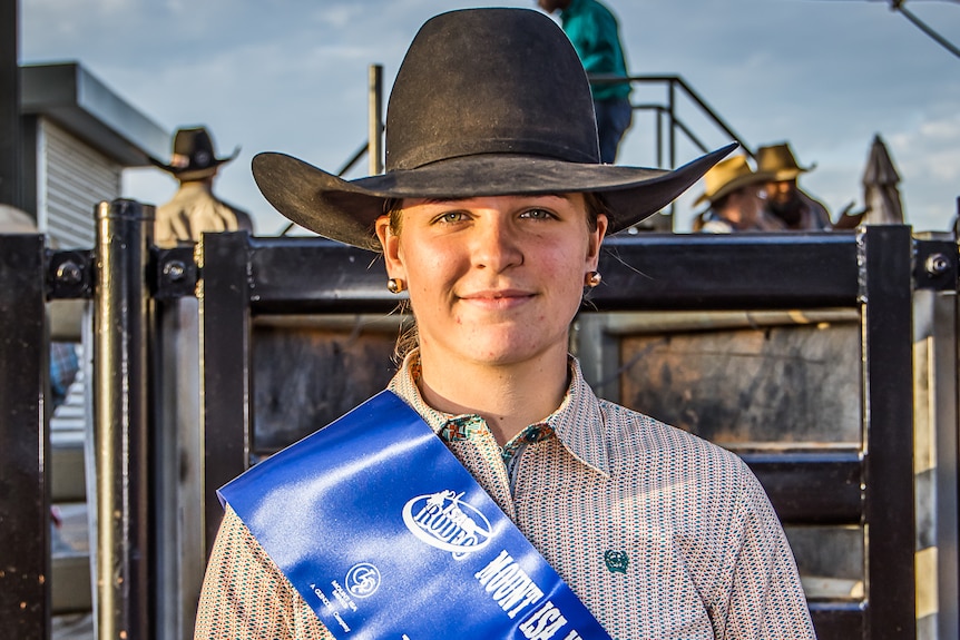 A cowgirl wears a blue sash and holds up a buckle prize at a rodeo