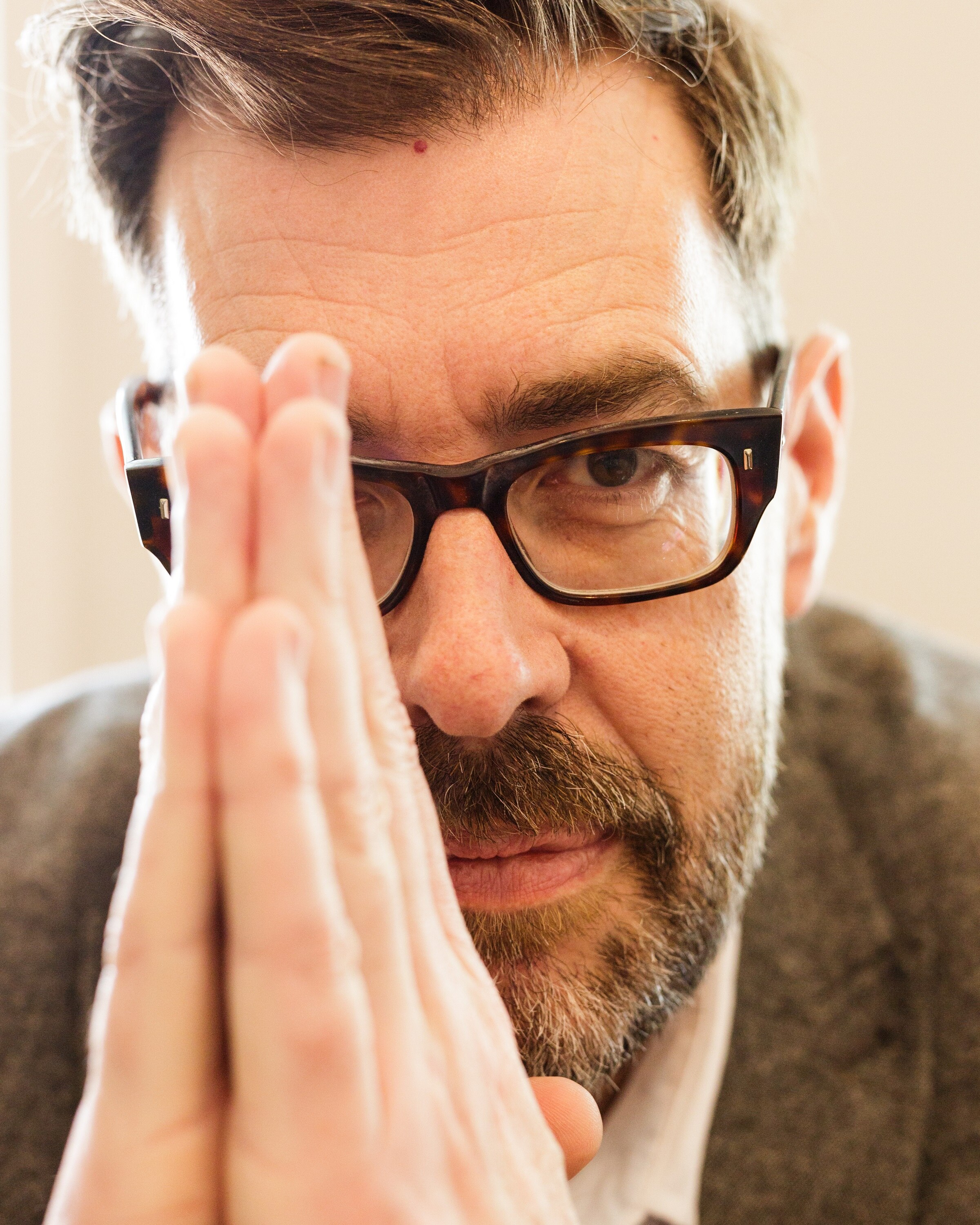 A close-up of a middle-aged white man with brown hair, a beard and dark-rimmed glasses holding his hands in front of his face