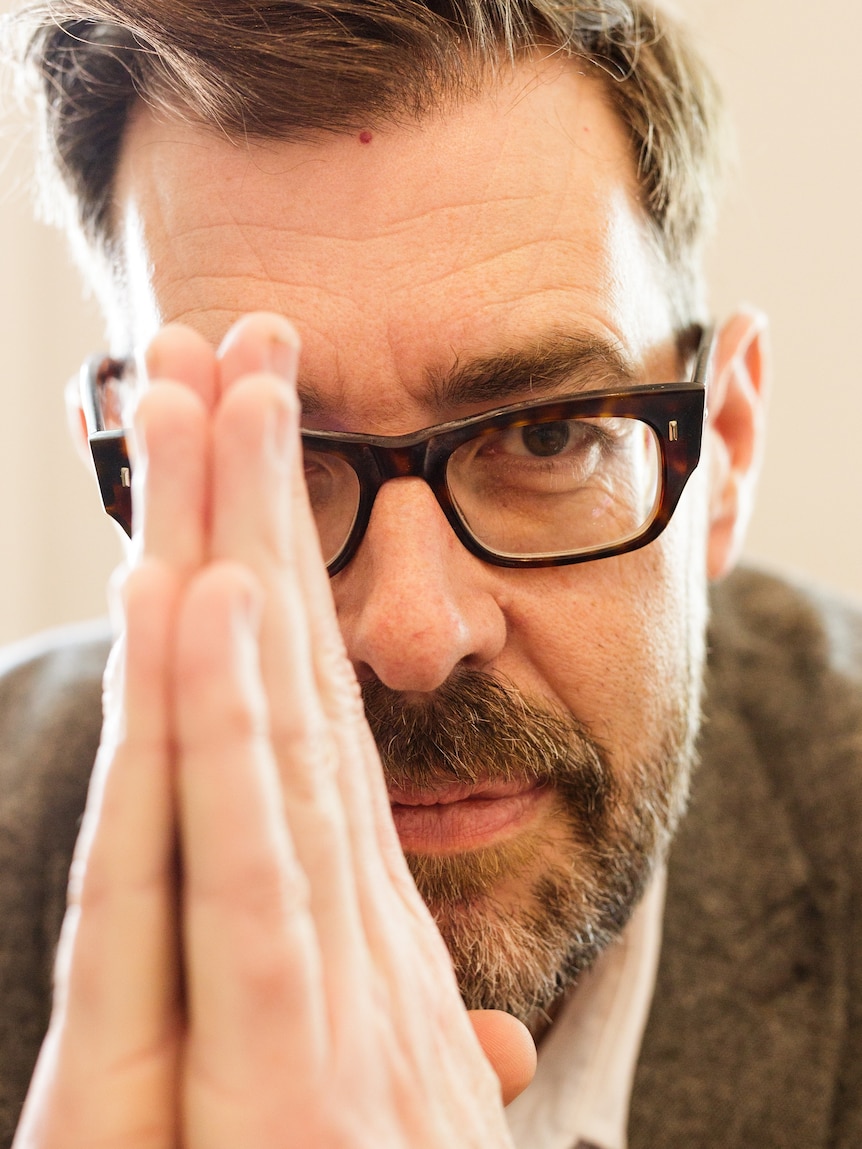 A close-up of a middle-aged white man with brown hair, a beard and dark-rimmed glasses holding his hands in front of his face