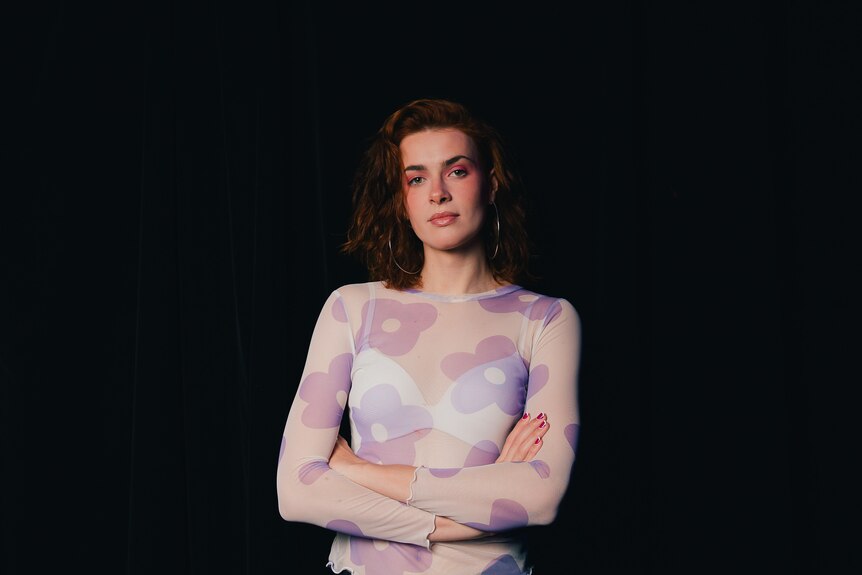 White trans woman with auburn shoulder-length hair wears floral pink turtle neck with arms folded in front of black curtain.