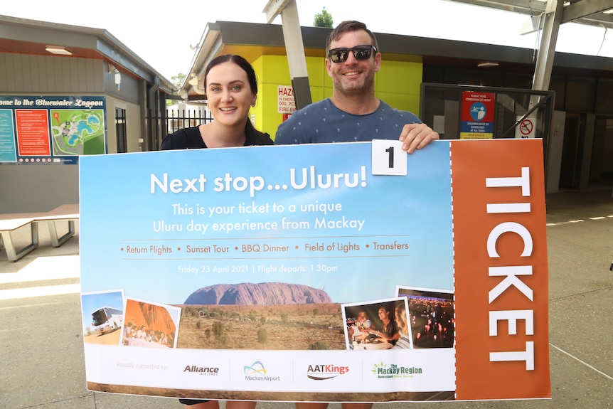 A male and female couple hold a large sign that says TICKET: Next stop Uluru
