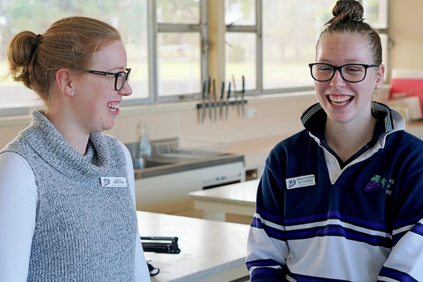Two young women wearing glasses share a laugh in a school classroom