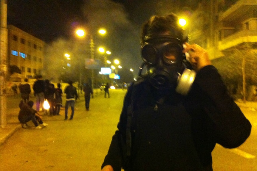 Brown wearing gas mask at night with rioters in background.
