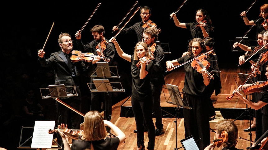 The Australian Chamber Orchestra stand performing on stage with bows raised dramatically in the air.