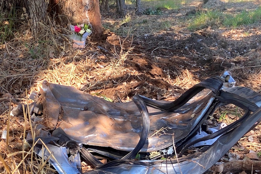 A box of flowers sits at the base of a tree in front of the wreckage of a car.