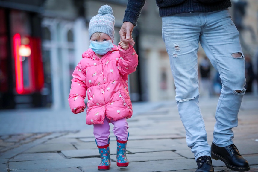 A small girl in a blue face mask, beanie and pink coat holds a man's hand 