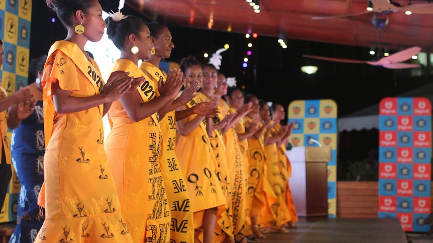Contestants for the Hiri Queen contest dressed in yellow, lined up on a stage and clapping.