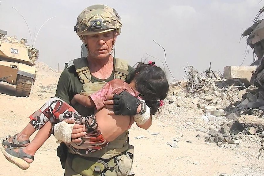 David Eubank, wearing body armour, carries a little girl through a rubble-lined street.