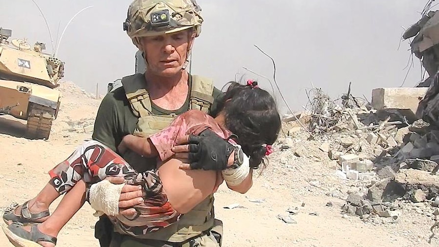 David Eubank, wearing body armour, carries a little girl through a rubble-lined street.