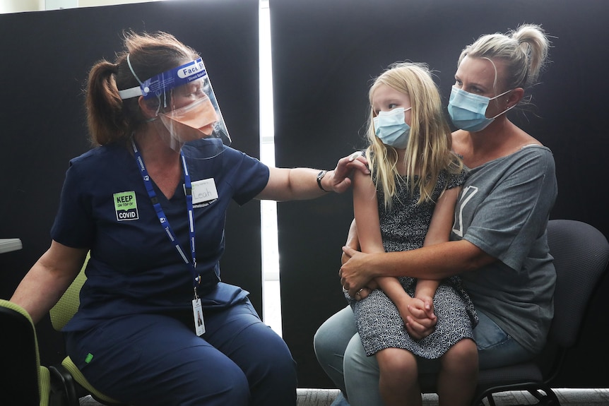 A young girl sits on her mum's knee as a nurse prepares to give her an injection