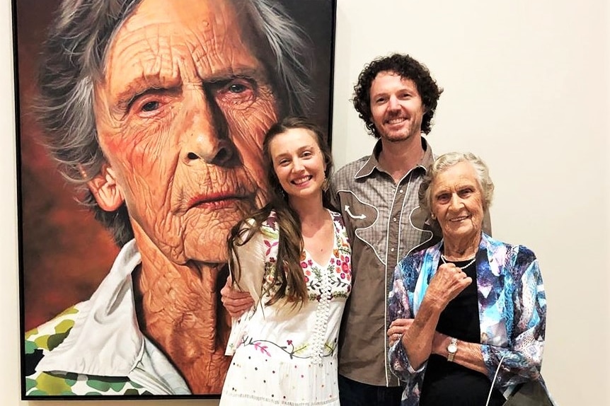 A young man and woman with elderly woman standing in front of portrait of elderly woman