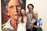 A young man and woman with elderly woman standing in front of portrait of elderly woman