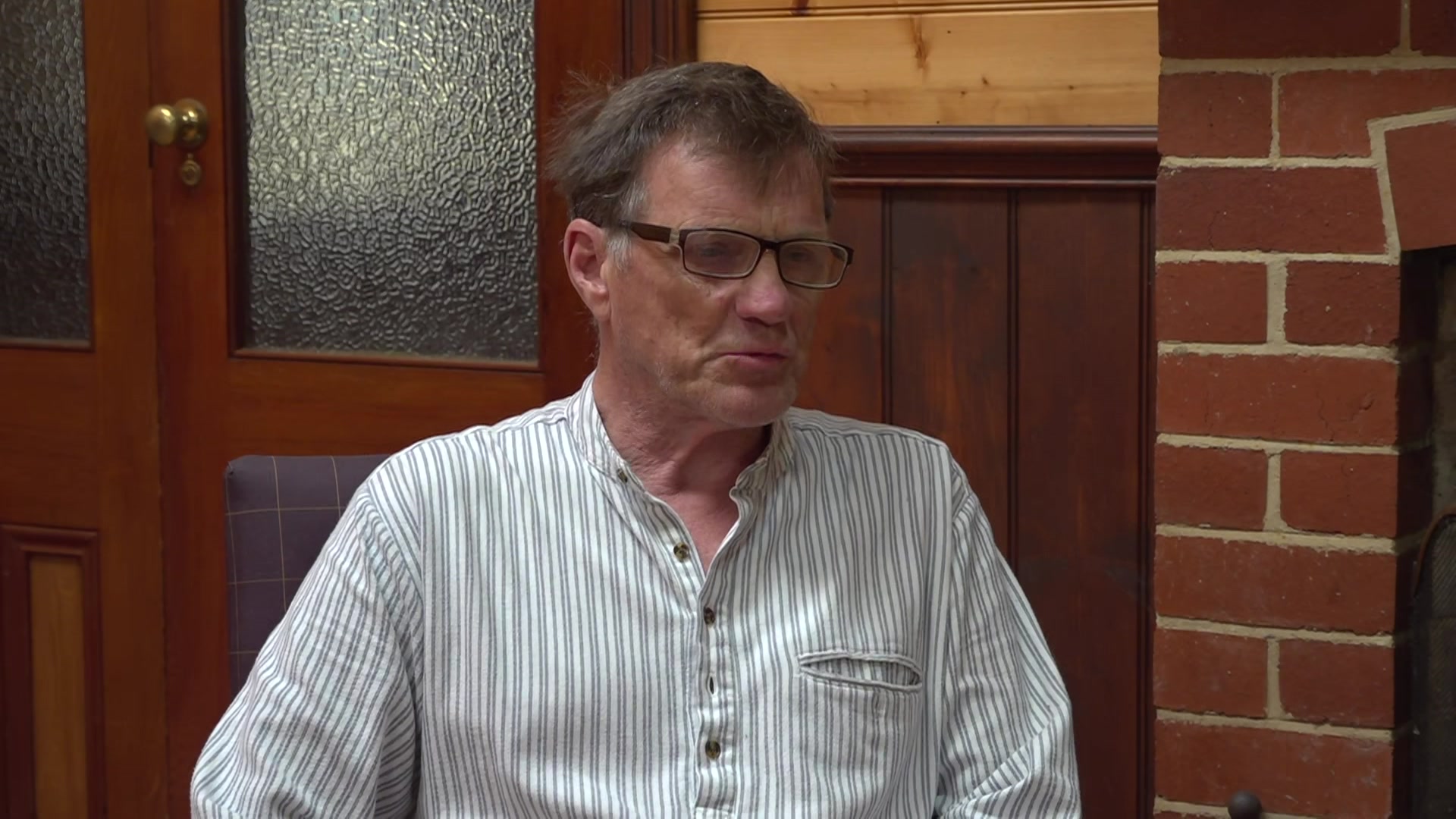 An elderly man in a striped business shirt and glasses sits in a chair, talking, in a brick and wood panelled room