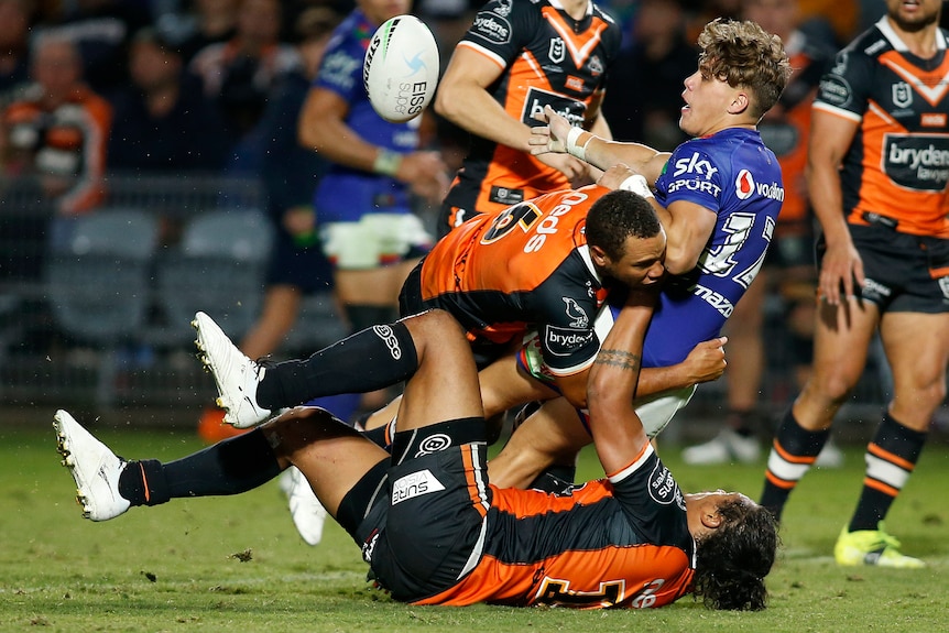 A Warriors NRL player offloads a pass while being tackled by two Wests Tigers' opponents.