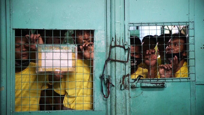 Prisoners lean against a metal door, their faces and hands poking through barred windows.