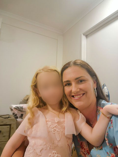 Uma mulher e sua filha, cujo rosto está desfocado, ficam lado a lado e sorriem.