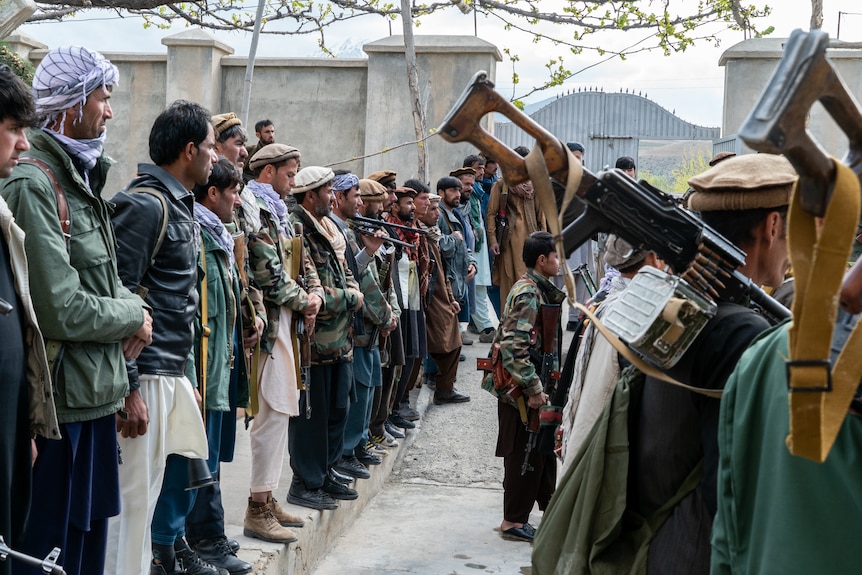 Men line up with guns over their shoulders and long robes listening to someone talk