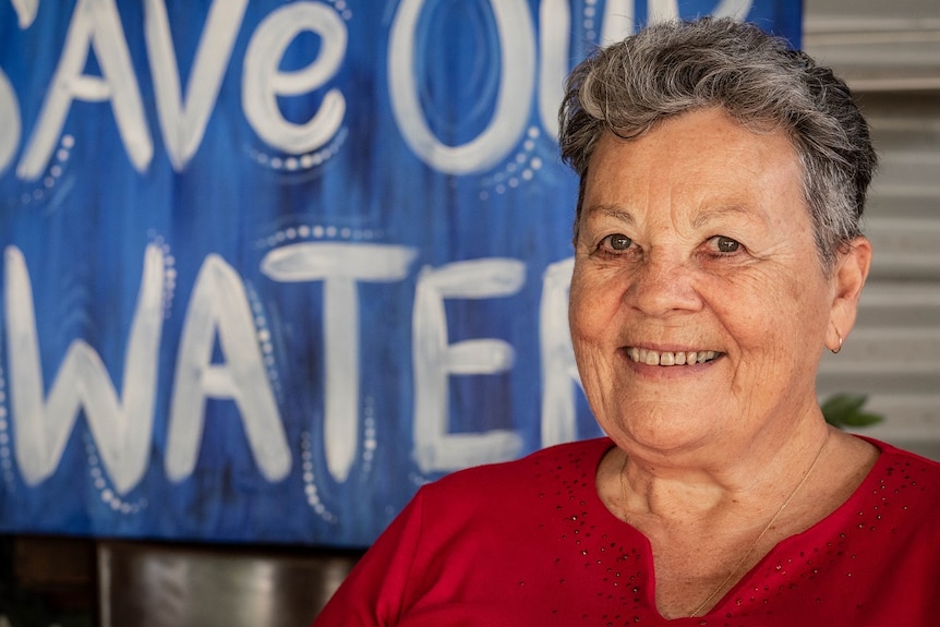 A smiling woman with short grey hair in the fore ground, a big sign reading save our water is behind her.
