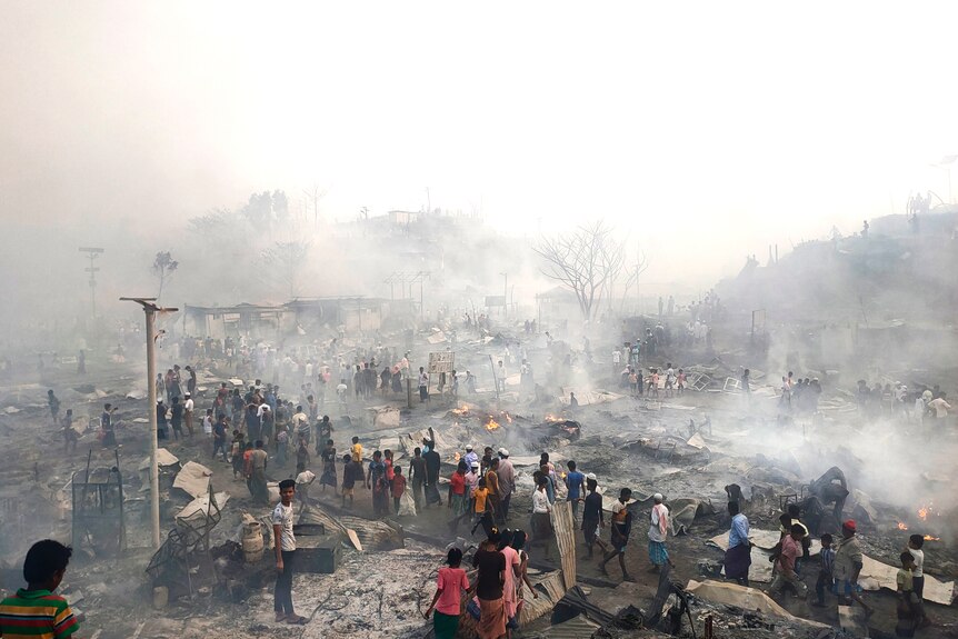Refugees walk through the charred and still smoking remains of homes after a massive fire.