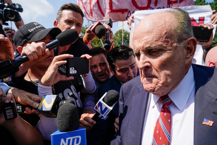 Rudy Giuliani, wearing a red tie and USA pin on his lapel, frowns as reporters shove microphones towards him
