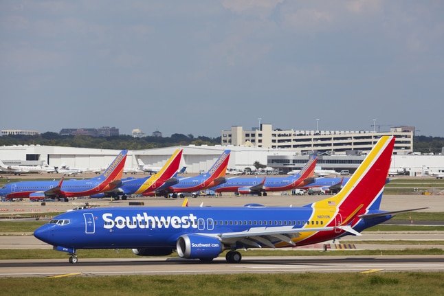 A plane is shown taxiing down a runway past a fleet of similar aircraft that are parked in the background.