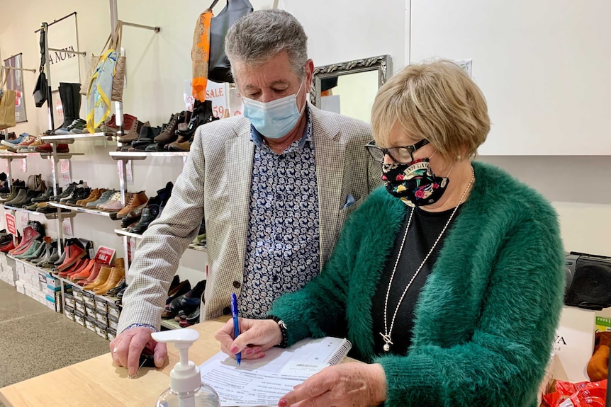 A man and woman wear face masks and stand behind the counter of a shoe store.