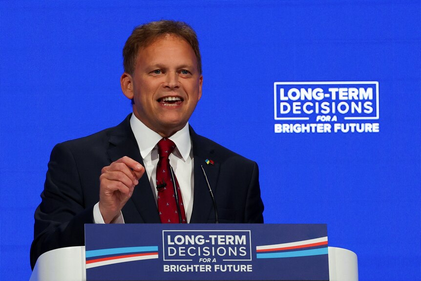 A happy-looking middle-aged white man in a suit gestures as he speaks behind a lectern in front of a blue background.