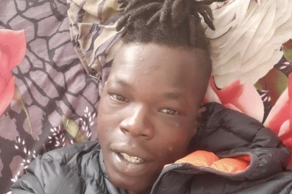 A selfie photo of young, dark man with dreads tied up on his head. He is lying down on a brightly coloured sheet or cover. 