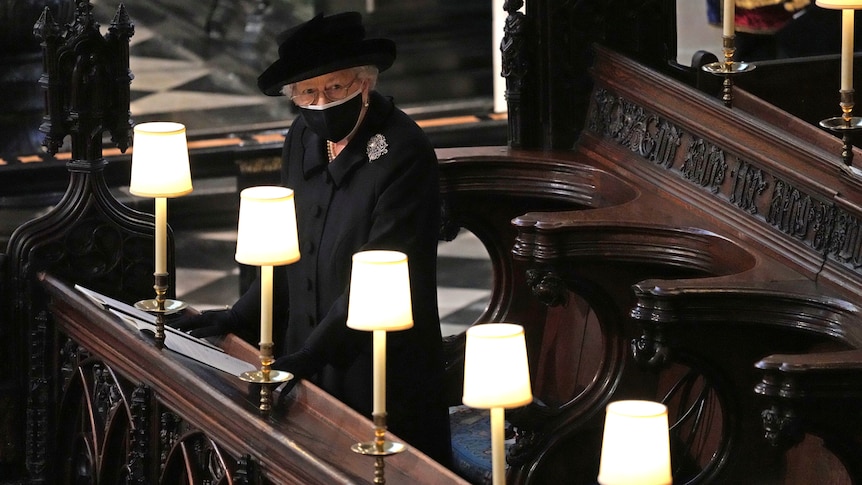 Wearing all black and a face mask, the Queen stands alone in the pews