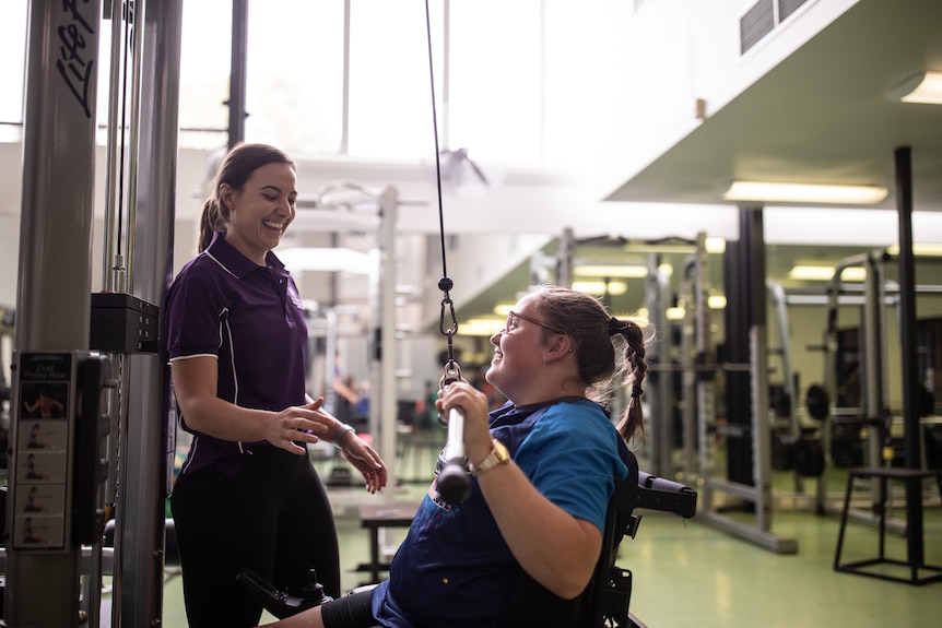 Chloe Tookey working out on a weight machine and smiling. Bridget Dodds is laughing and assisting. 