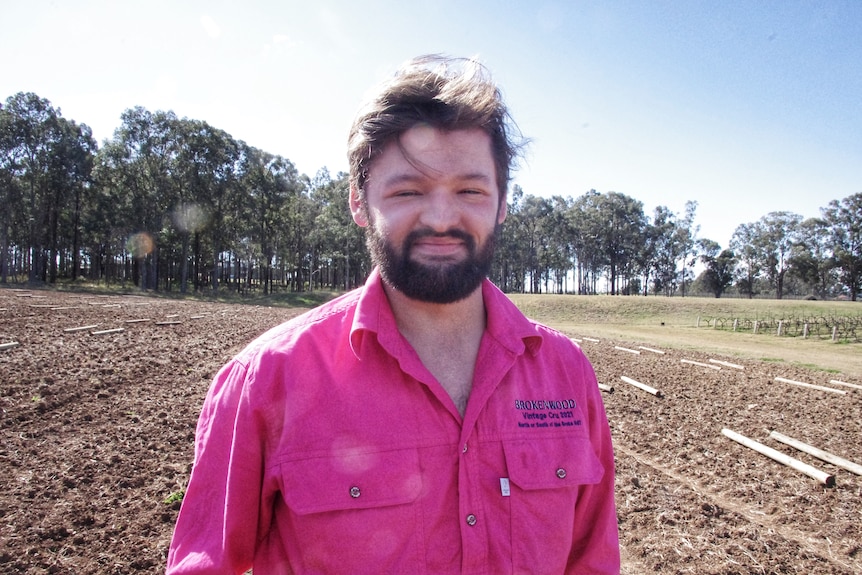 A young man in a pink shirt stands in a barren vineyard.