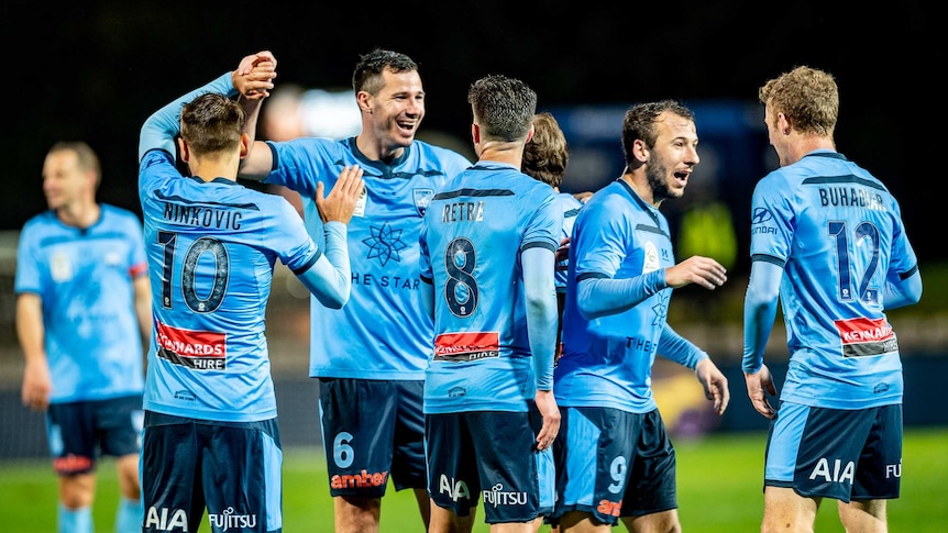 Sydney FC players celebrate together in pale blue shirts on a football pitch