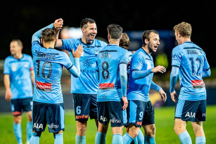 Sydney FC players celebrate together in pale blue shirts on a football pitch