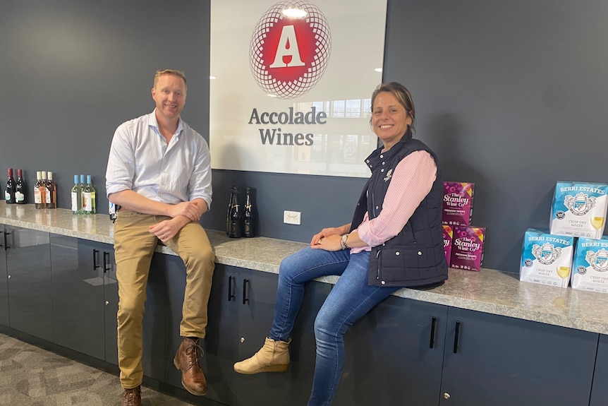 A man and a woman sit on a bench with casks and bottles of wine around them. 