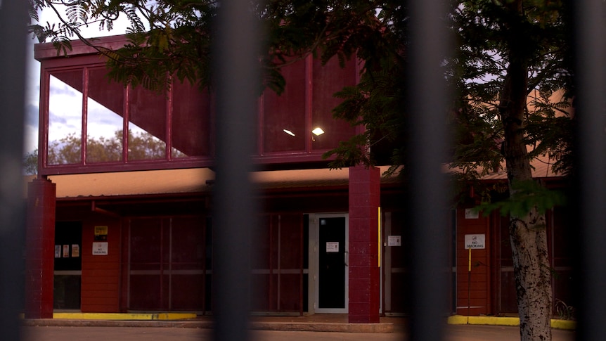A maroon single story hospital building can be seen through the bars of the fence surrounding it.