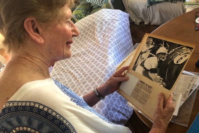 An older woman in a white shirt looking at a black and white photograph of herself wearing a jockey's outfit and riding a horse.