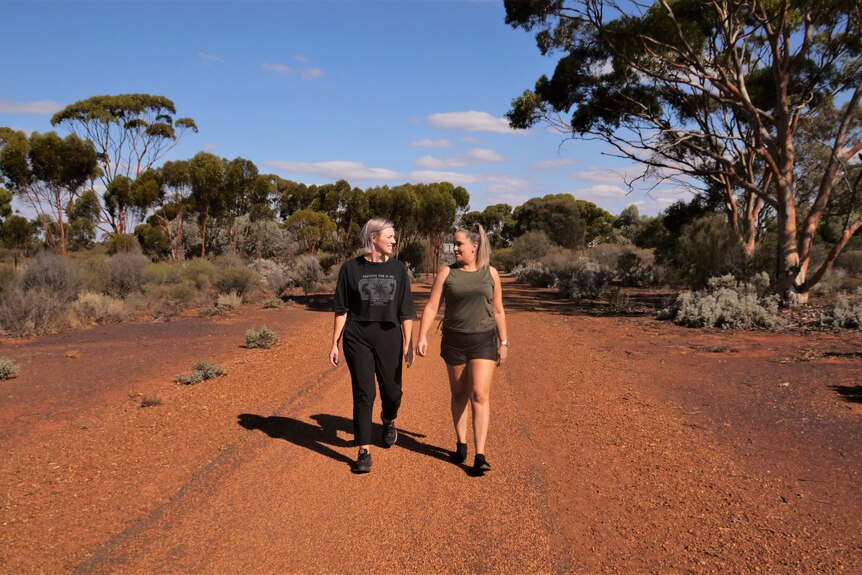 Two woman wearing activewear walk on a red sandy track surrounded by vivid blue sky and scrub and gum trees
