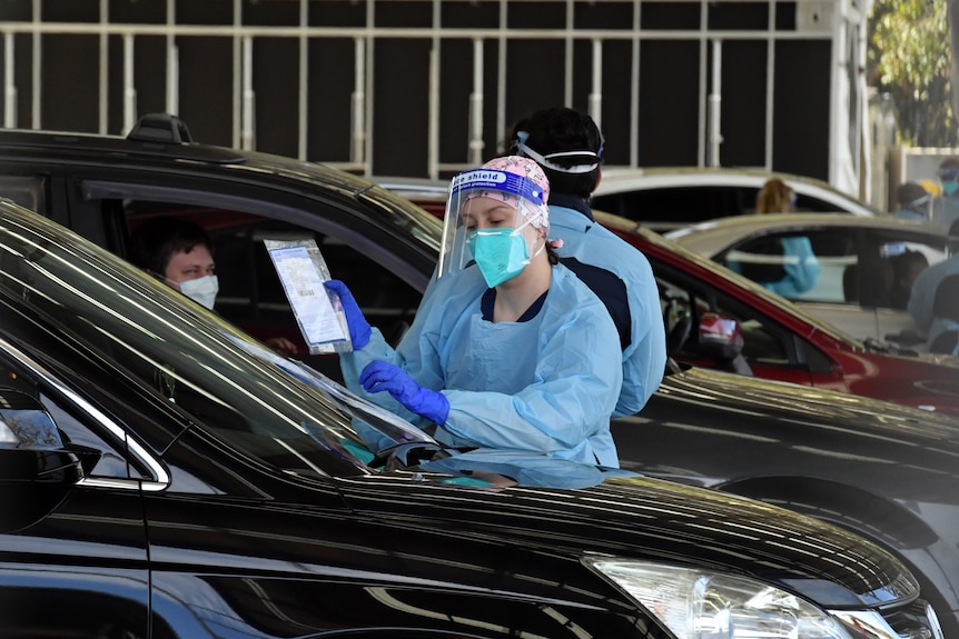 A woman wearing a medical gown, gloves and face mask and carrying a clipboard talks to a passenger in a car.
